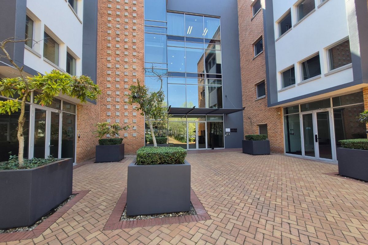 A modern commercial courtyard featuring brick and glass architecture, paved flooring, and integrated planters leading to a central glass-fronted building entrance.