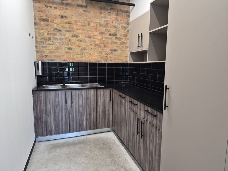 A modern scullery or kitchenette area featuring exposed brickwork, black subway tile backsplash, dark stone countertops, and contemporary wood-grain cabinetry with black hardware.