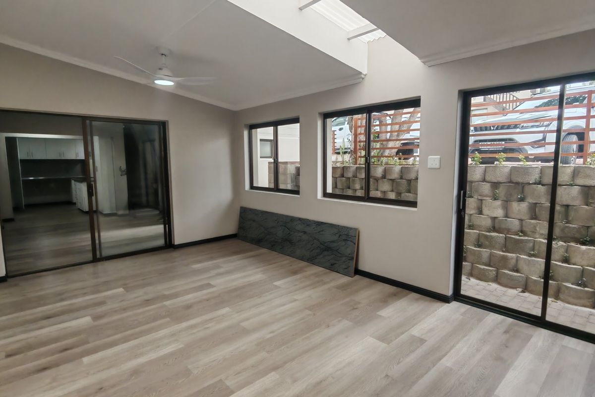 A bright, open-plan living area featuring modern laminate flooring, a ceiling fan, a skylight, and large sliding doors leading to an outdoor retaining wall.