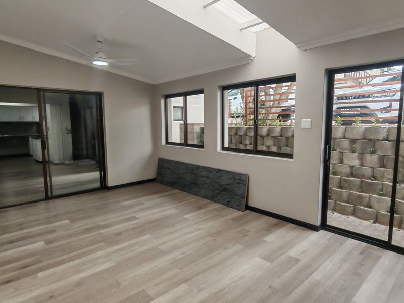 A bright, open-plan living area featuring modern laminate flooring, a ceiling fan, a skylight, and large sliding doors leading to an outdoor retaining wall.