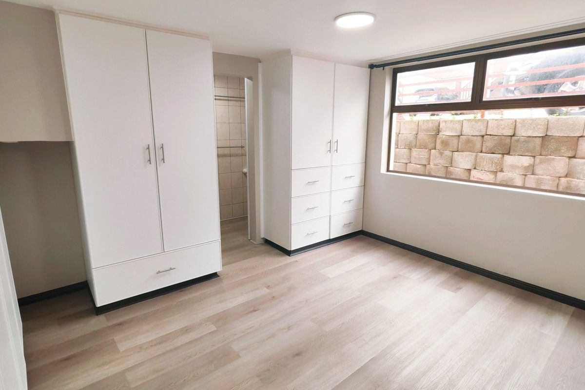 A bright, empty bedroom featuring modern laminate flooring, ample built-in cupboard storage, and a large window overlooking a retaining wall.