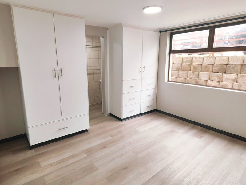 A bright, empty bedroom featuring modern laminate flooring, ample built-in cupboard storage, and a large window overlooking a retaining wall.