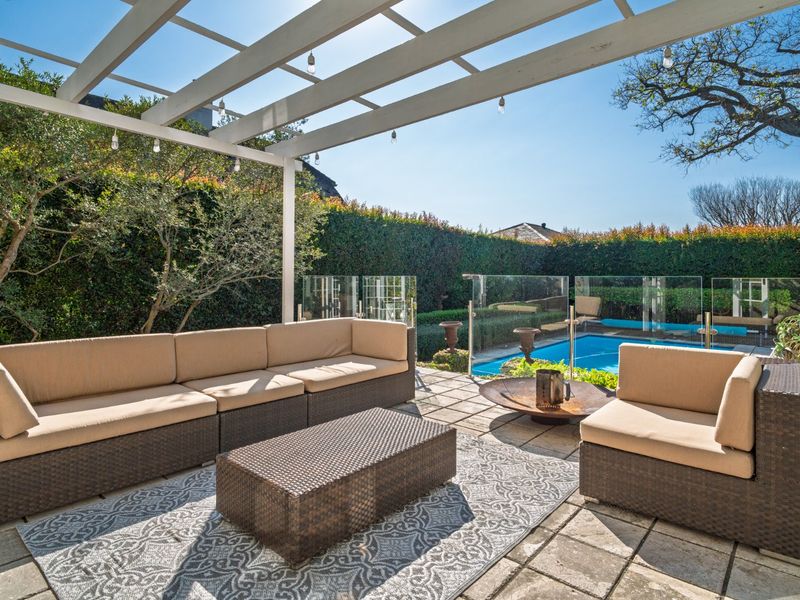 Outdoor patio area featuring a white pergola, paved flooring, and a glass-fenced swimming pool, perfect for entertaining.