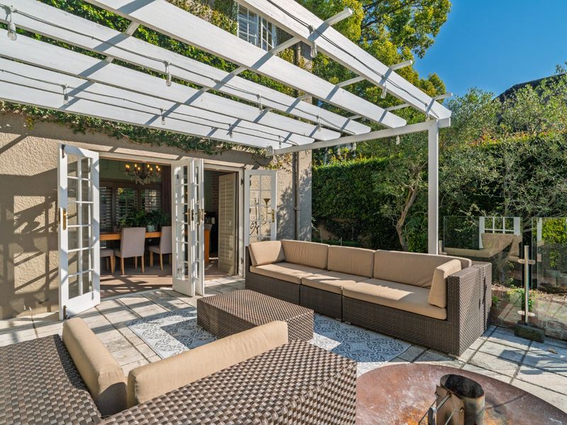 A well-maintained outdoor patio featuring a white pergola, paved flooring, and glass balustrades, providing seamless access to the interior living space through french doors.