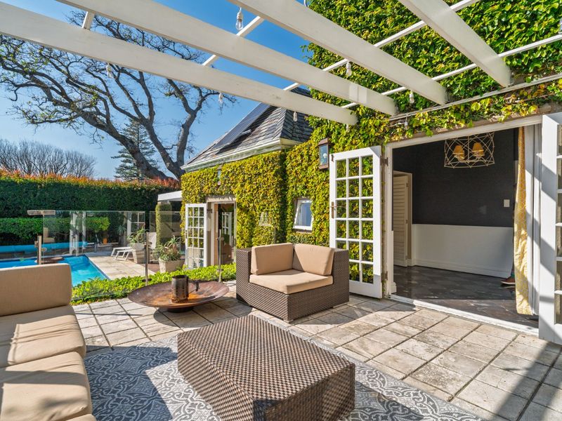 A covered outdoor patio featuring stone paving, a white pergola, and glass balustrades, providing seamless access to the pool area and garden.