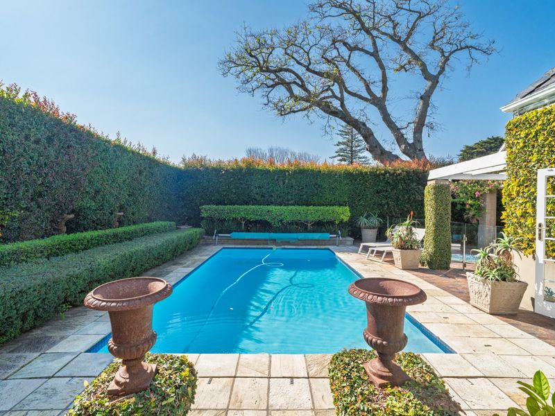 A well-maintained outdoor swimming pool area featuring stone paving, manicured hedges, and glass safety fencing, providing a private and serene garden setting.