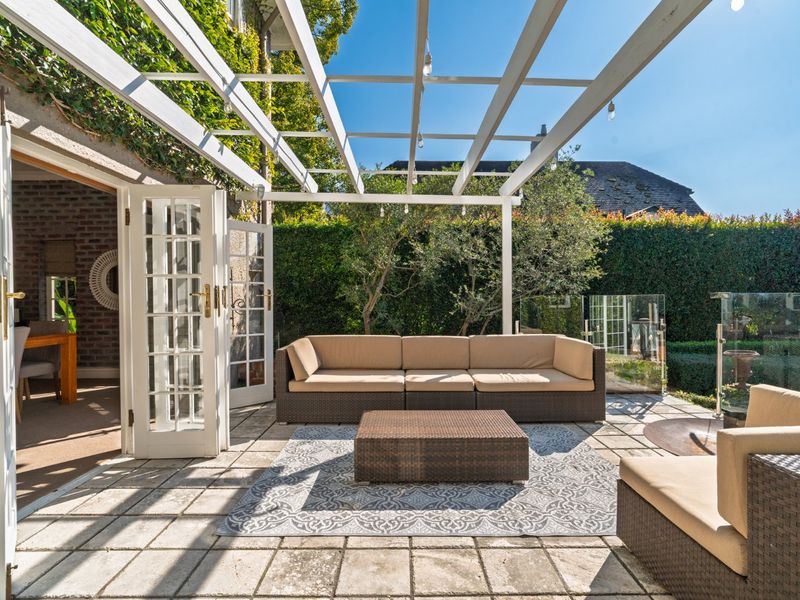 A well-maintained outdoor patio featuring a white pergola, paved flooring, and glass balustrades, providing a seamless transition from the interior living space.