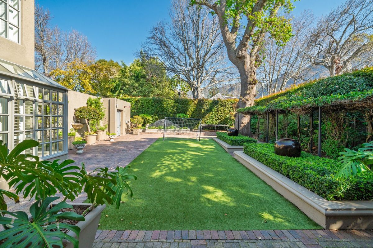 A well-maintained rear garden featuring a central artificial turf lawn, brick paved patio area, raised garden beds, and established perimeter hedging.