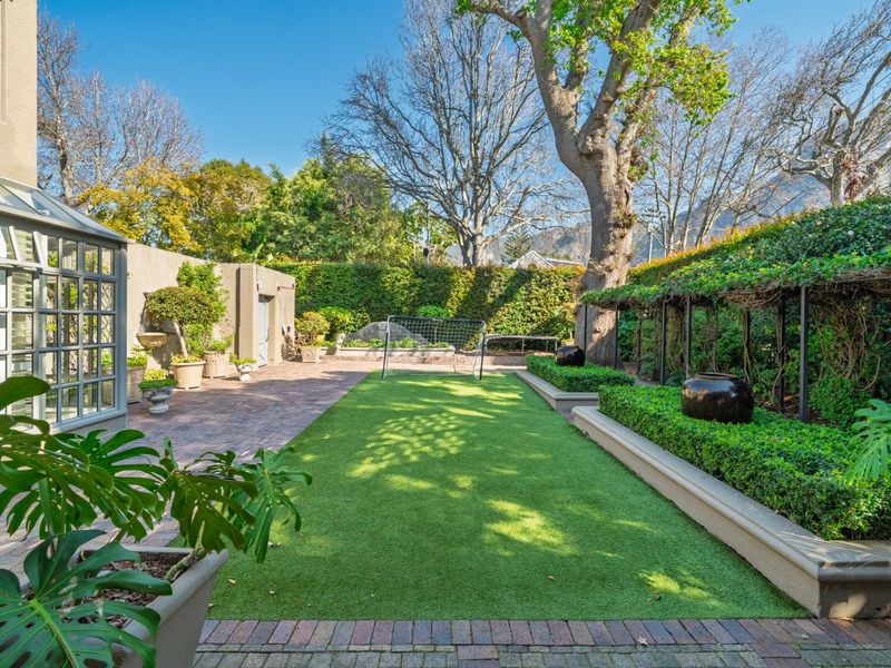A well-maintained rear garden featuring a central artificial turf lawn, brick paved patio area, raised garden beds, and established perimeter hedging.