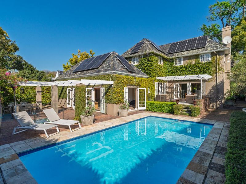 A beautifully landscaped rear garden featuring a sparkling swimming pool, stone-paved patio, pergola, and a charming ivy-clad home with solar panels.