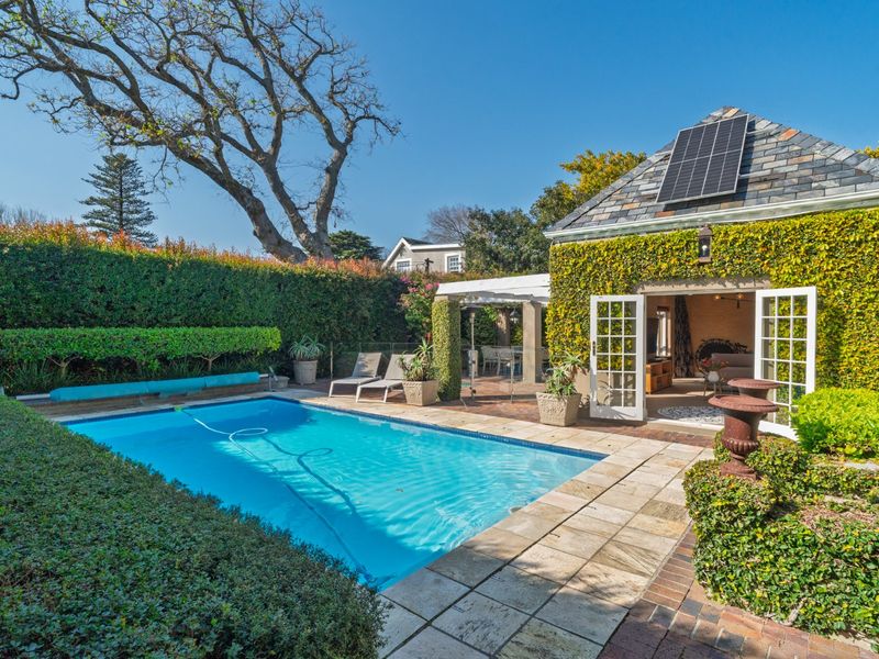 A well-maintained outdoor pool area featuring stone paving, lush hedging, and a charming outbuilding with solar panels and French doors.