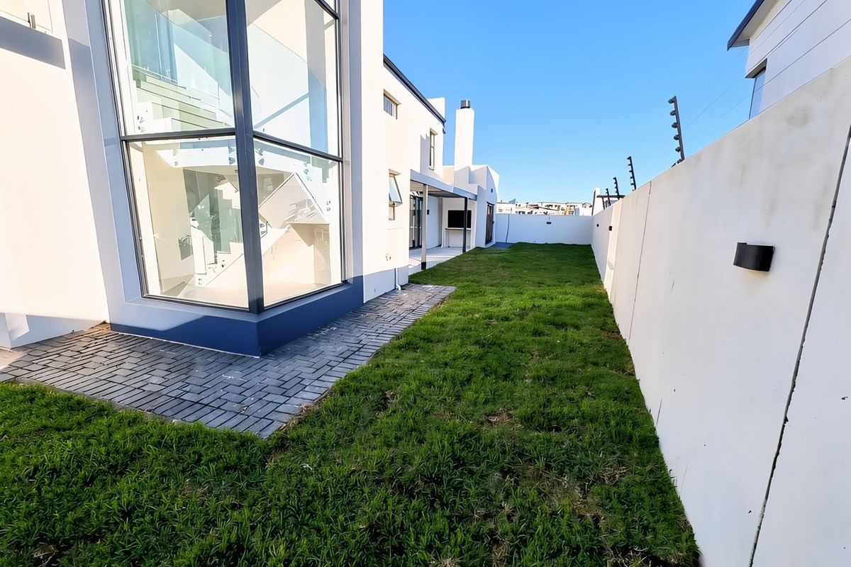 Rear garden area featuring a modern double-volume glass facade, paved walkway, lawn, and boundary wall with integrated electric fencing.
