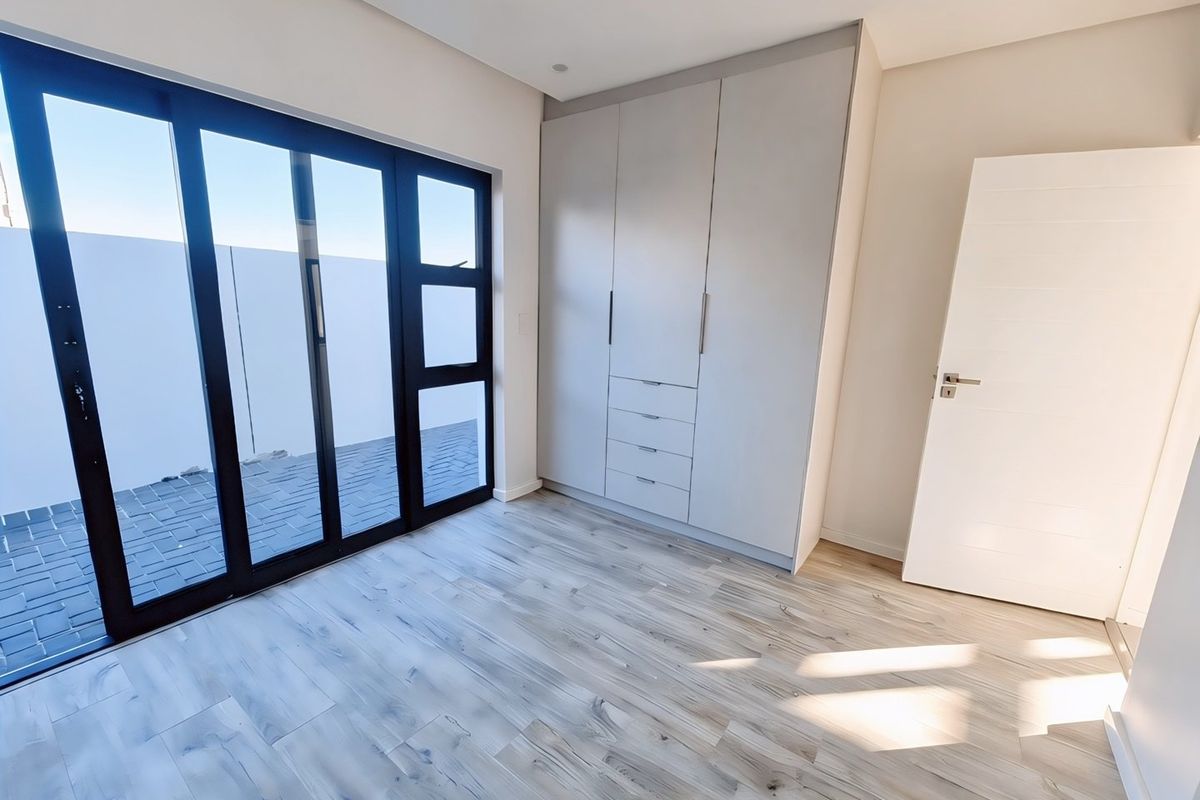 A bright, modern bedroom featuring light-toned laminate flooring, built-in cupboards, and a large sliding glass door providing access to the exterior.