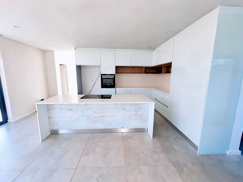 Modern, minimalist kitchen featuring a central island with integrated cooktop, sleek white cabinetry, built-in oven, and large format floor tiles.