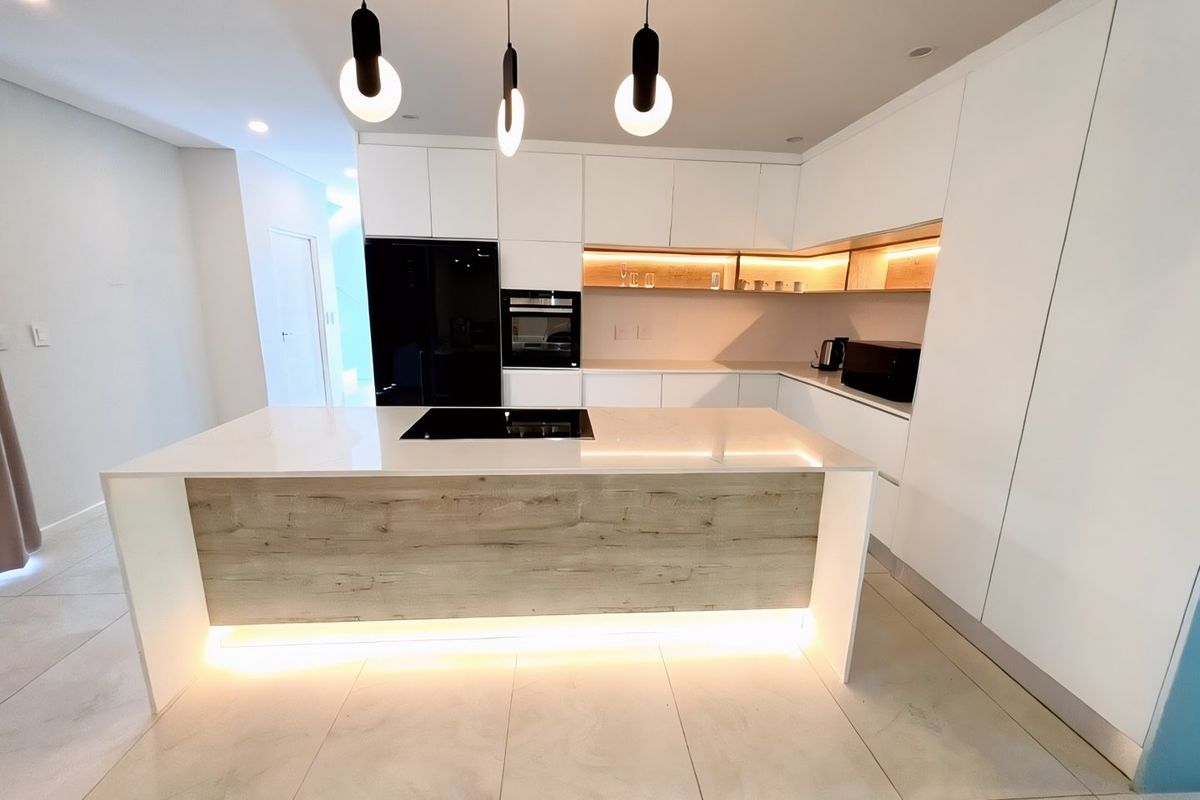 Modern, minimalist kitchen featuring a central island with induction cooktop, sleek white cabinetry, integrated appliances, and warm under-cabinet LED lighting.