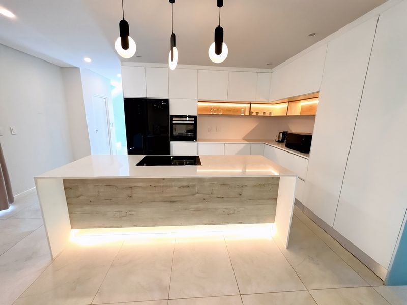 Modern, minimalist kitchen featuring a central island with induction cooktop, sleek white cabinetry, integrated appliances, and warm under-cabinet LED lighting.