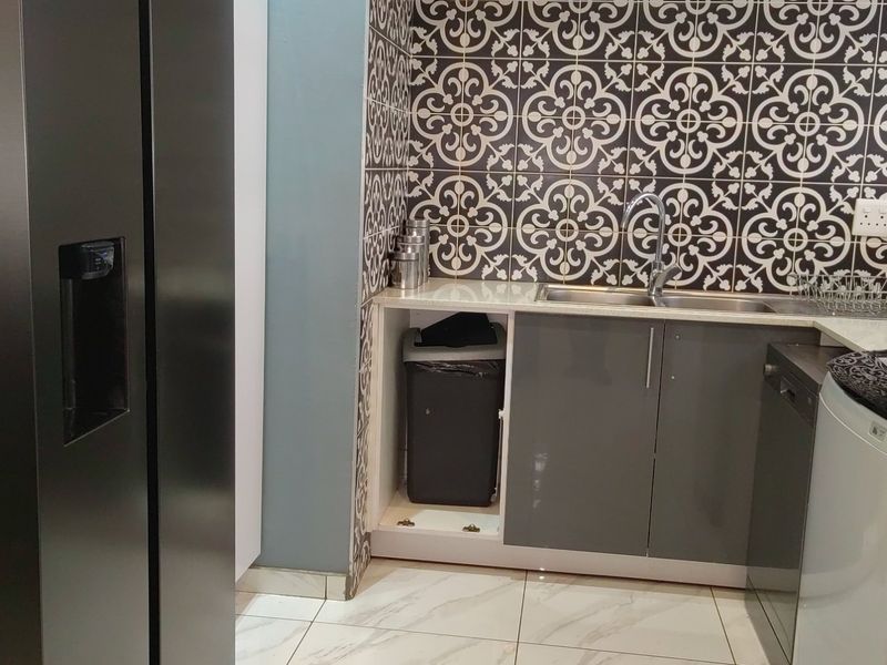 A functional scullery area featuring patterned wall tiles, grey cabinetry, a stainless steel sink, and tiled flooring.