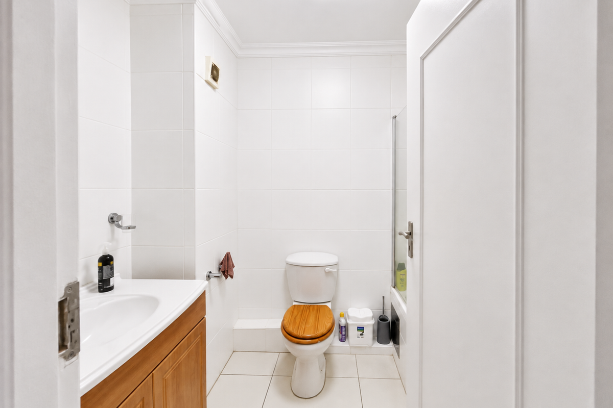 A clean, functional bathroom featuring white wall tiles, a wooden vanity cabinet, a ceramic sink, and a shower enclosure.