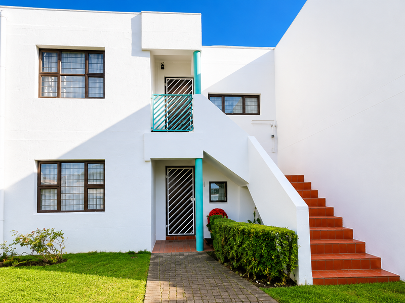 A well-maintained apartment exterior featuring white rendered walls, a tiled staircase, security gates, and a paved walkway leading to the entrance.
