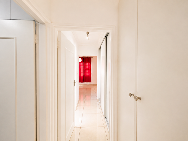 A bright, clean hallway featuring neutral tiled flooring and ample built-in cupboard storage, leading towards an adjacent room with red window treatments.