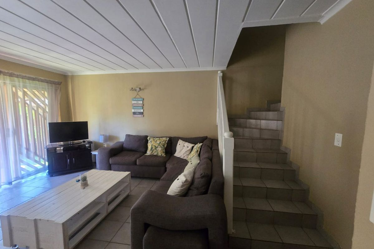 A living room featuring tiled flooring, a staircase with a white balustrade, and a timber panelled ceiling, with access to an outdoor area.