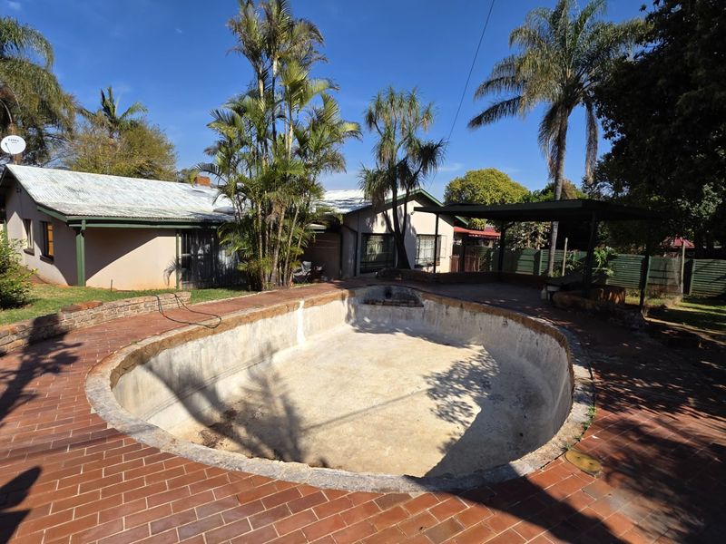 Outdoor pool area featuring a large empty swimming pool surrounded by brick paving and a covered patio structure.