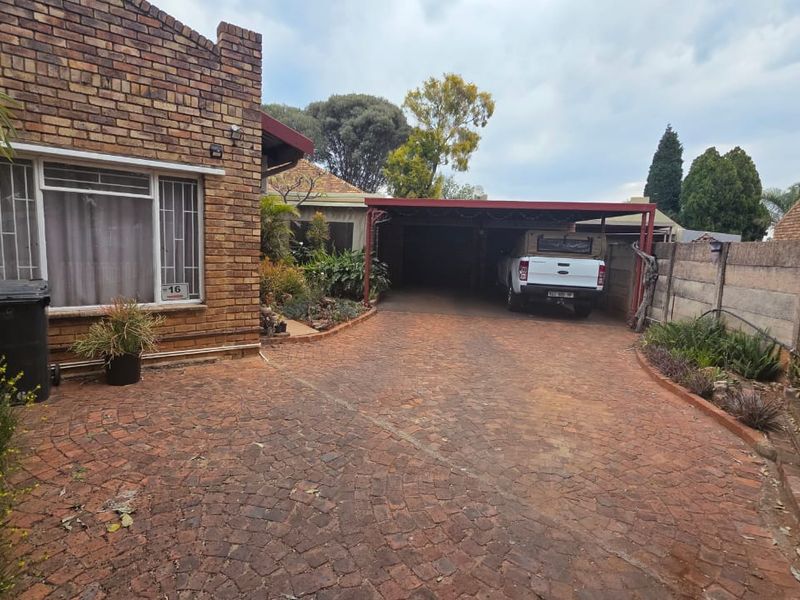 Front exterior view featuring a brick facade, paved driveway, and a covered carport area with a concrete boundary wall.