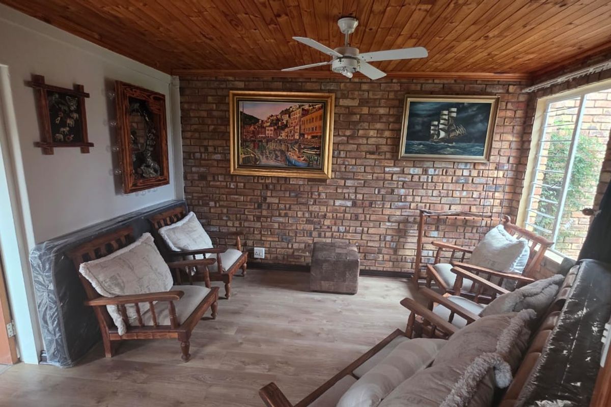 A cozy living room featuring an exposed brick feature wall, timber ceiling, ceiling fan, and laminate flooring with natural light from a large window.