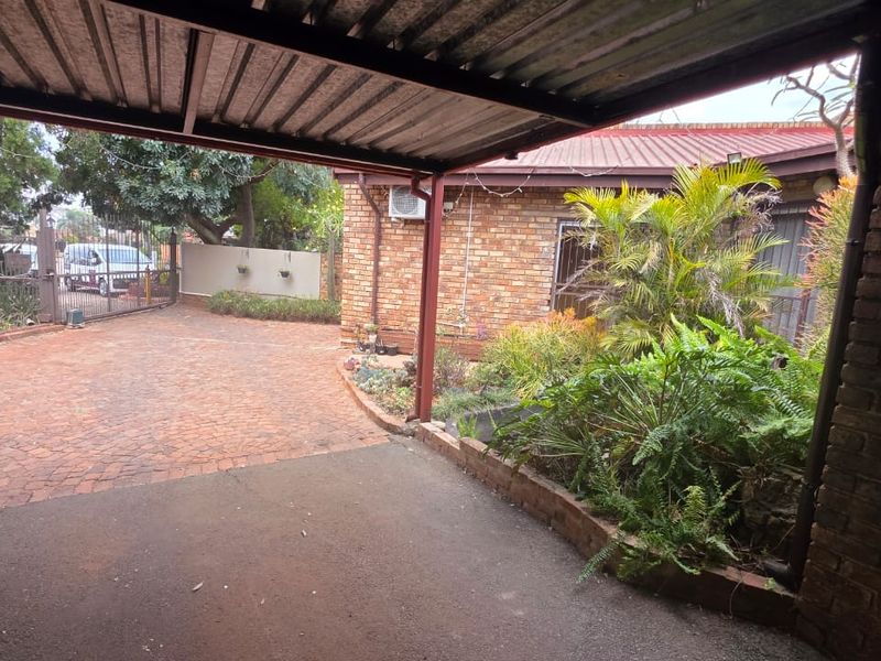 A view of the front exterior featuring a covered carport, brick driveway, and established garden beds against a facebrick facade.