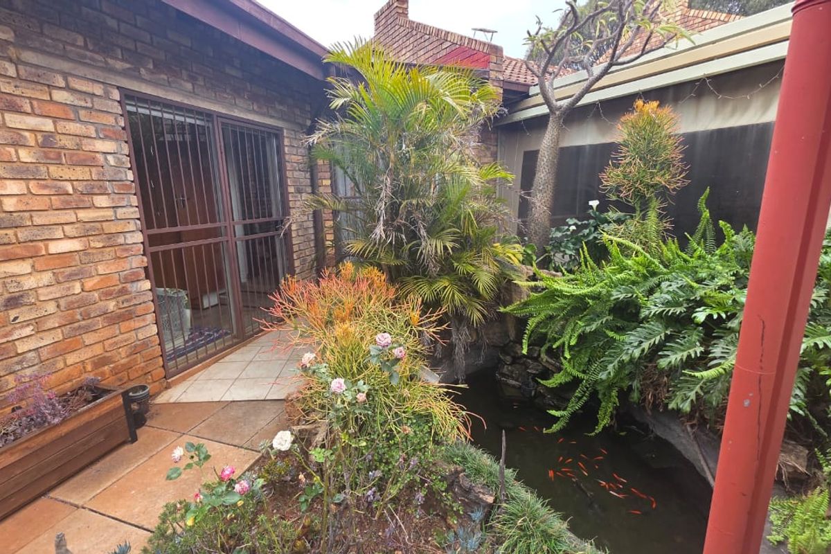 A rear courtyard area featuring a brick facade, tiled patio access, and a built-in fish pond surrounded by established garden landscaping.