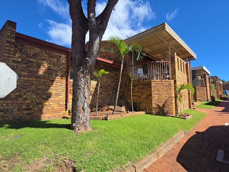 A view of the townhouse exterior featuring face-brick construction, a covered patio area, and a well-maintained front garden with paved driveway access.