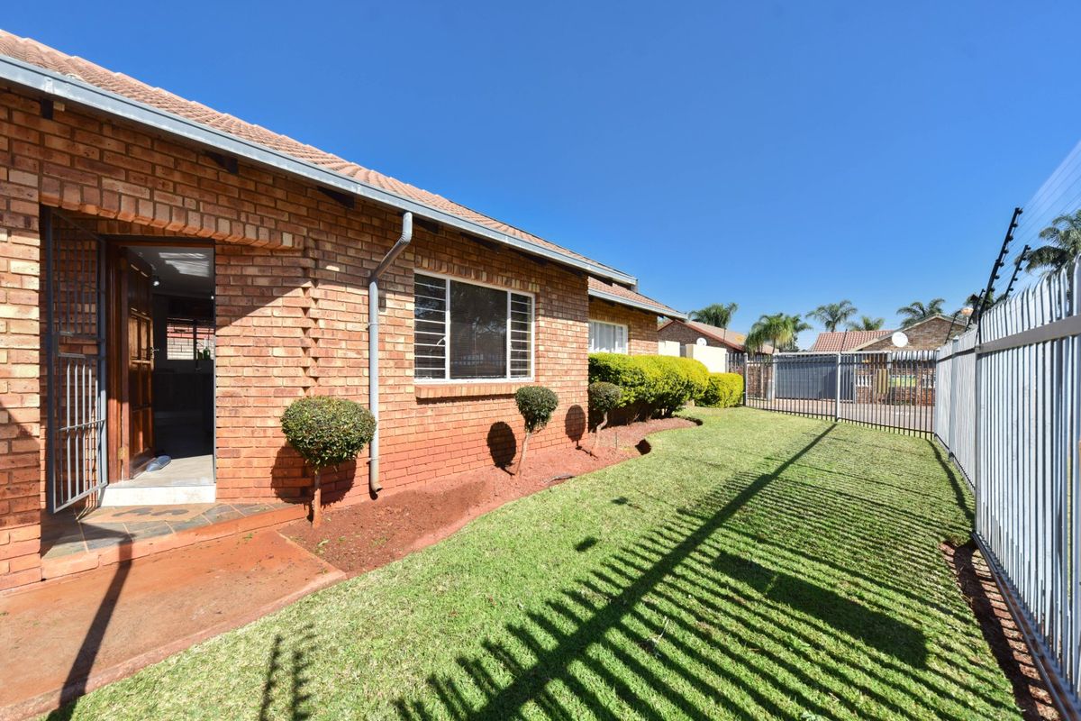 A well-maintained rear exterior view of a face brick house featuring a manicured lawn, secure perimeter fencing, and electric security wiring.