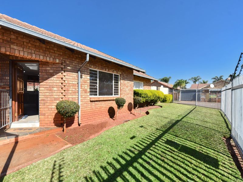 A well-maintained rear exterior view of a face brick house featuring a manicured lawn, secure perimeter fencing, and electric security wiring.