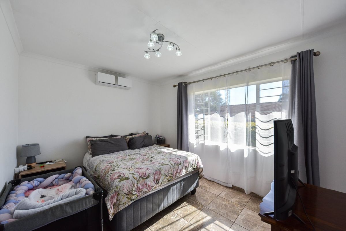 A functional bedroom featuring tiled flooring, a wall-mounted air conditioning unit, and a window with security bars, providing natural light.