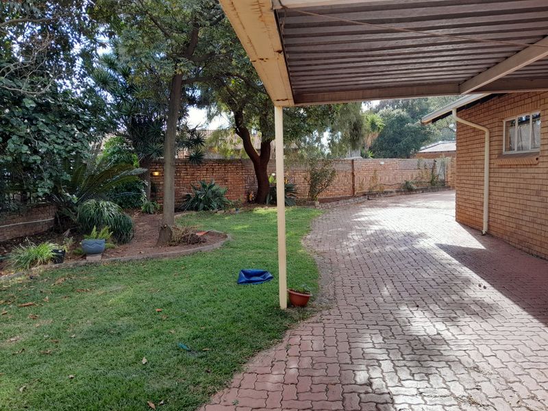 A view of the front exterior featuring a paved driveway, a covered carport structure, and a landscaped garden area with a brick boundary wall.