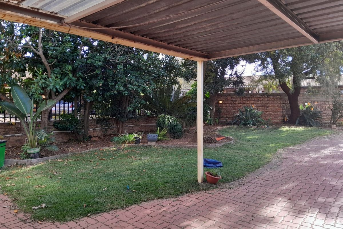 A rear garden area featuring a paved driveway, a lawn, and a covered carport structure with a brick boundary wall.