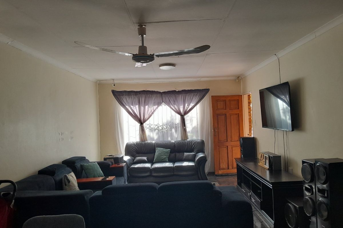 A living room featuring tiled flooring, a ceiling fan, and a wooden entrance door, showing signs of wear on the walls and ceiling.