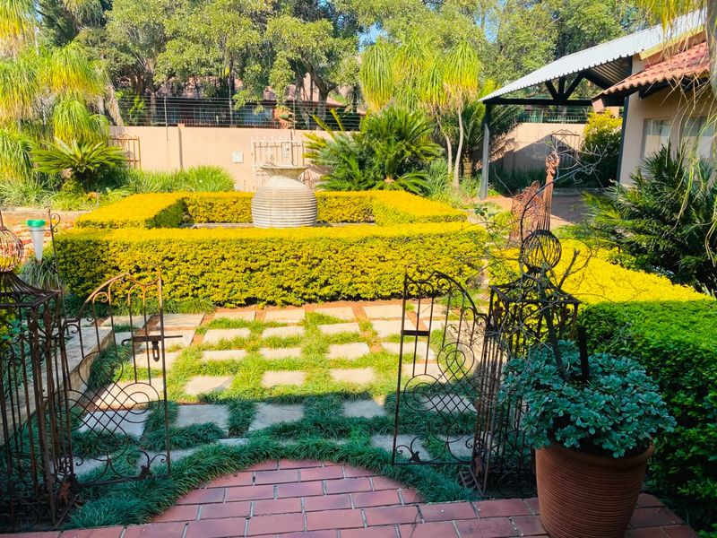 A well-maintained garden featuring manicured hedges, stone paving, a central water feature, and a wrought iron gate leading to a covered patio area.
