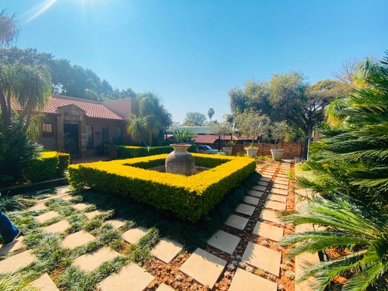 A well-maintained garden featuring a central stone fountain, manicured hedges, and stone paved pathways leading toward the main house entrance.