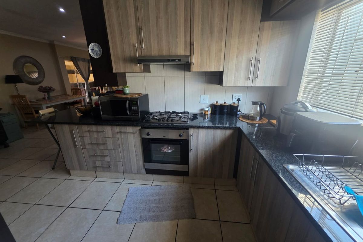 A functional kitchen featuring wood-look cabinetry, granite countertops, a gas hob, built-in oven, and tiled flooring, with an adjacent dining area visible.