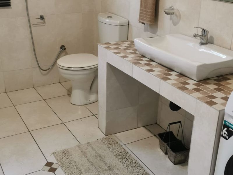 A functional bathroom featuring a tiled vanity with a vessel sink, wall-mounted mirror, toilet, and bidet spray, finished with neutral ceramic wall and floor tiles.