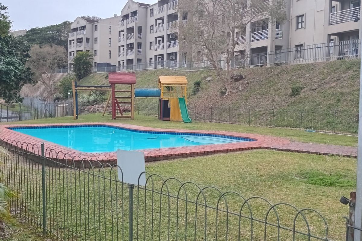 Communal apartment complex pool area featuring a swimming pool with brick coping, metal fencing, and a children's play structure on the lawn.