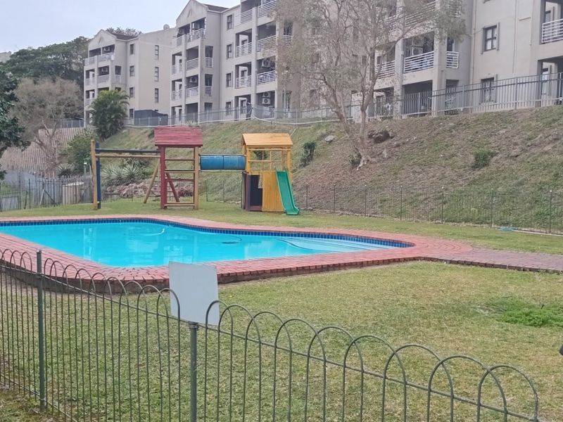 Communal apartment complex pool area featuring a swimming pool with brick coping, metal fencing, and a children's play structure on the lawn.