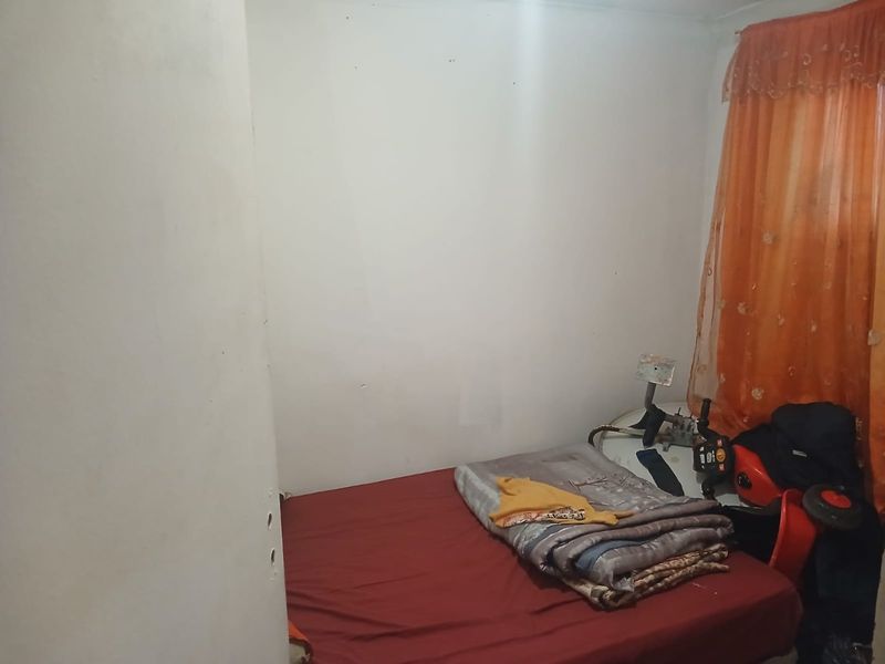 A compact bedroom featuring plain walls and a window with orange curtains, requiring cosmetic updates to improve its overall appeal.