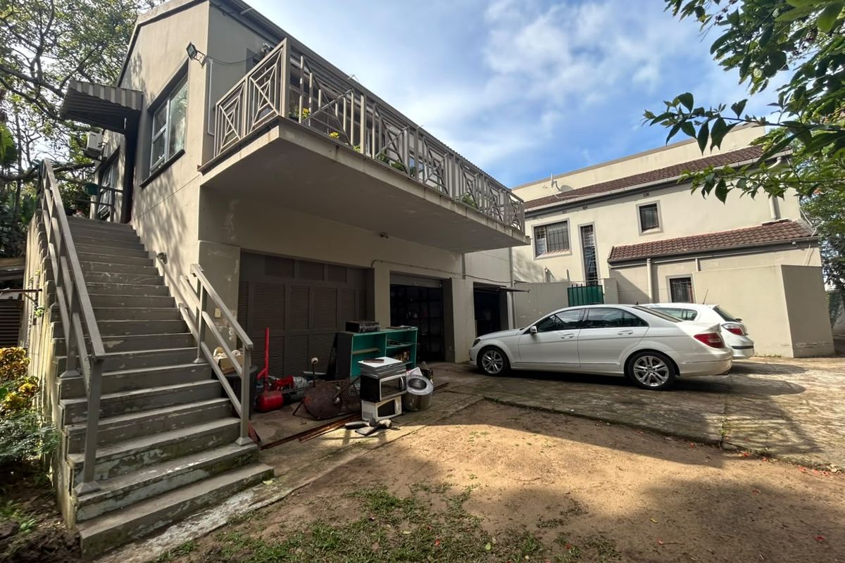 Rear exterior view of the property featuring a raised balcony with metal railings, external staircase, and ground-level garage access with a paved driveway area.