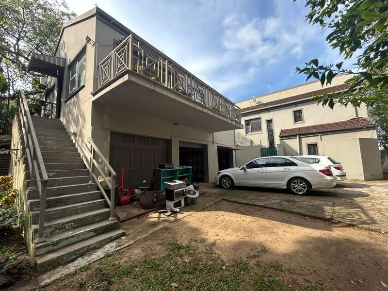 Rear exterior view of the property featuring a raised balcony with metal railings, external staircase, and ground-level garage access with a paved driveway area.