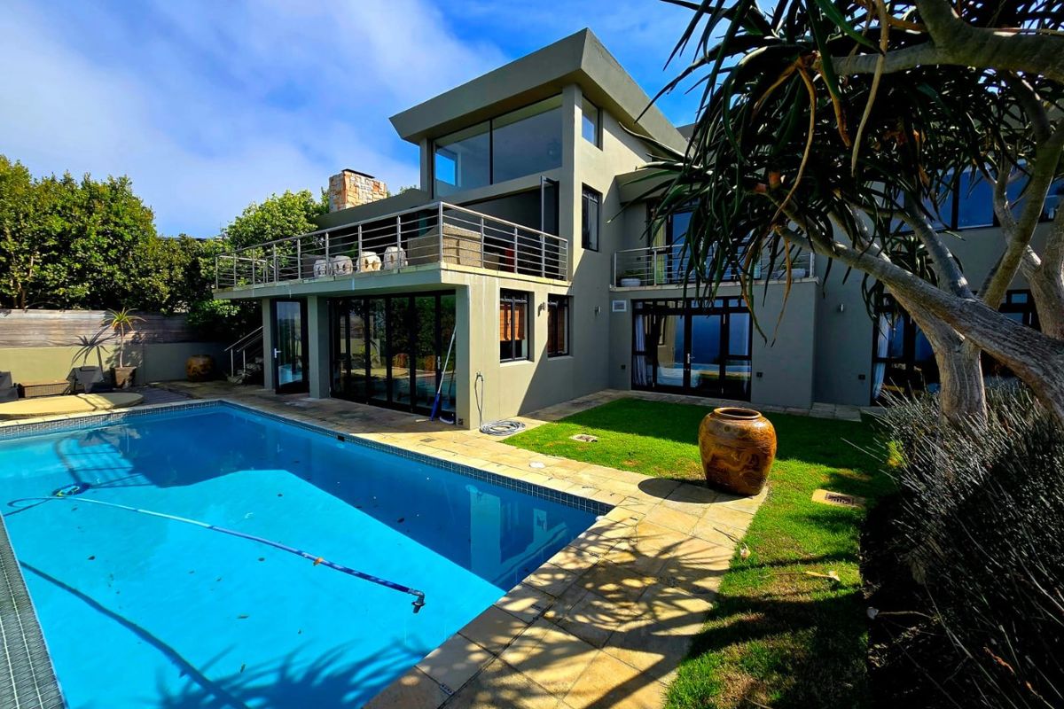 Rear view of a modern multi-story house featuring a swimming pool, stone-paved patio, and a spacious upper-level balcony with stainless steel railings.