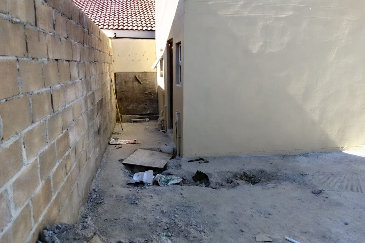 A rear exterior area showing an unfinished ground surface, exposed plumbing, and a concrete block boundary wall in need of landscaping and maintenance.