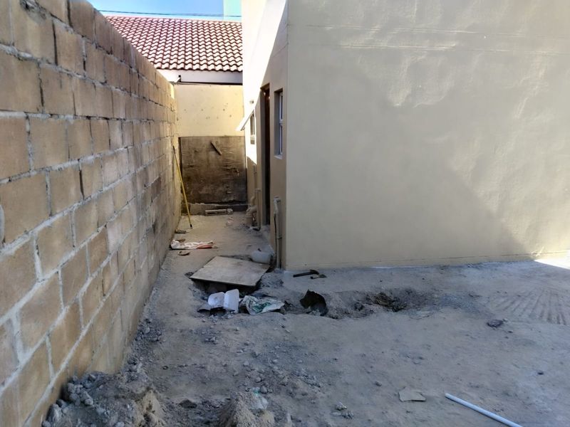 A rear exterior area showing an unfinished ground surface, exposed plumbing, and a concrete block boundary wall in need of landscaping and maintenance.