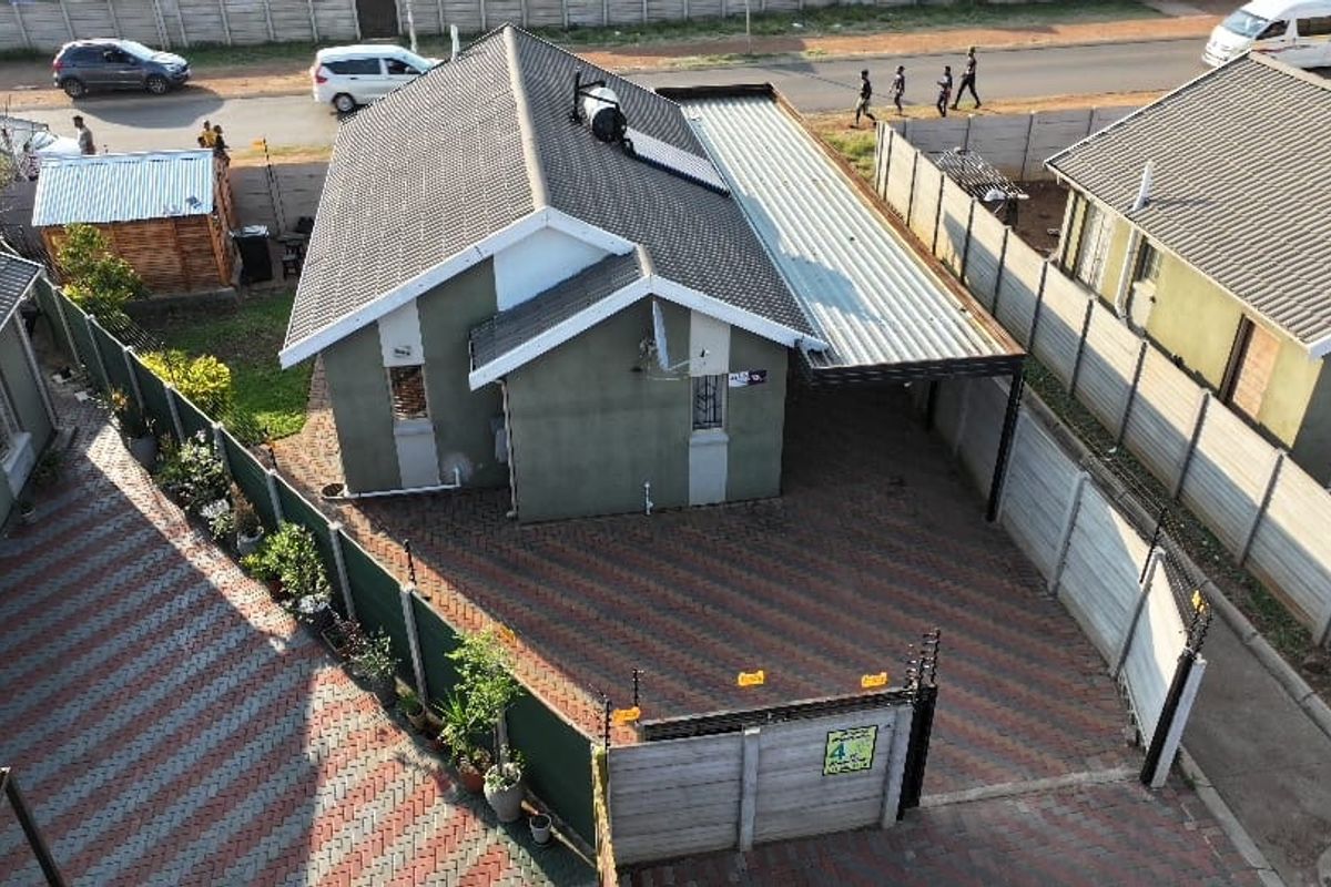Aerial view of a residential property featuring a tiled roof with solar geyser, paved driveway, carport, and secure perimeter walling with electric fencing.